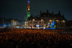A huge crowd fills Bradford Centenary Square at night - the buildings around it are lite up in blues and greens.